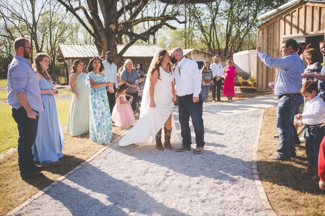 Bride and groom share a kiss on the ceremony path — the bride wearing decorated cowboy boots beneath her dress